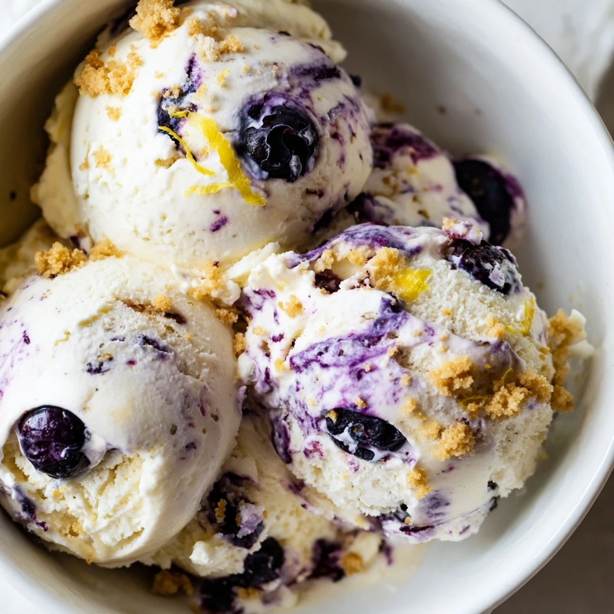 A scoop of creamy Lemon Blueberry Cheesecake Ice Cream reveals blueberry swirls and graham cracker crumbs in a chilled bowl.