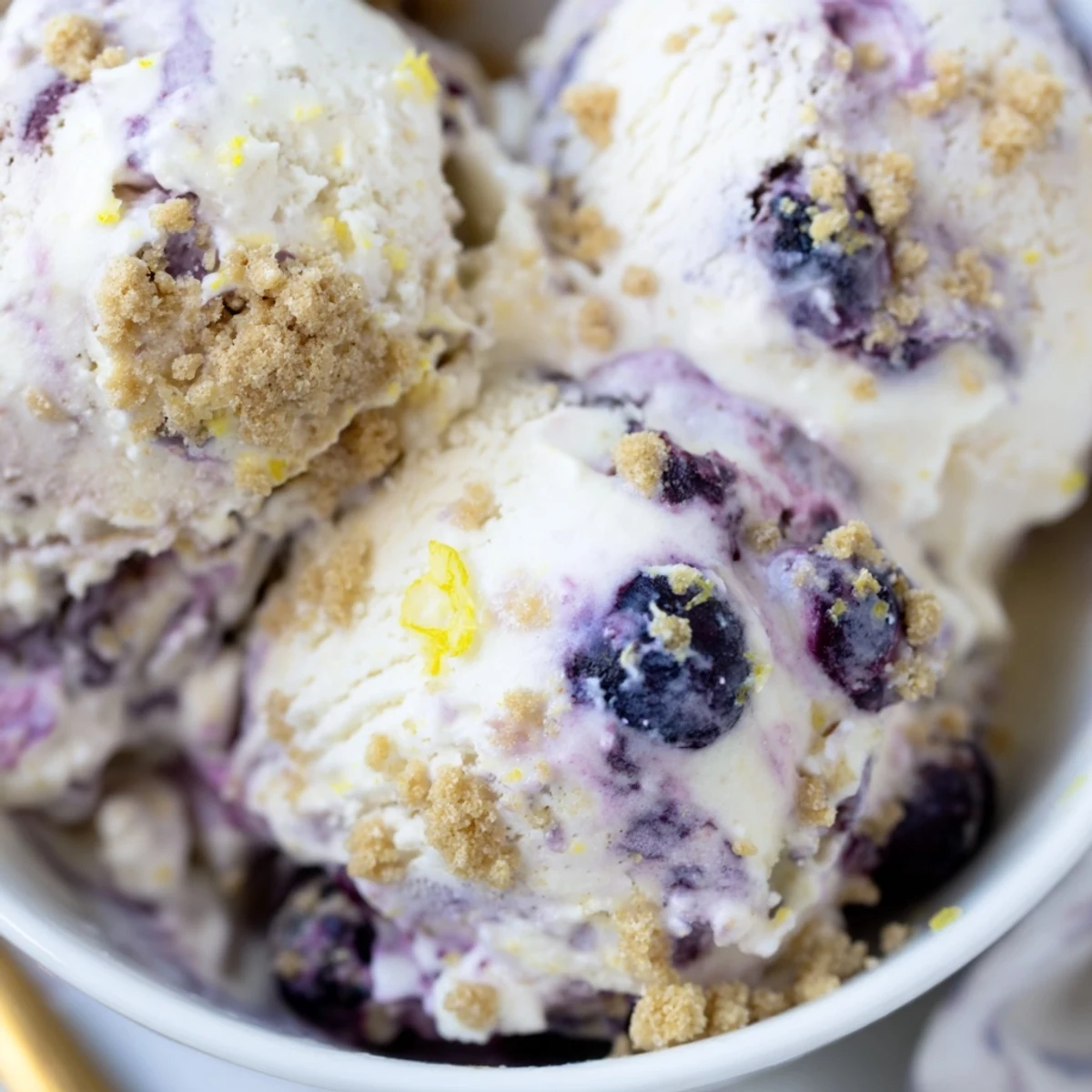Close up shot of tangy Lemon Blueberry Cheesecake Ice Cream in a waffle cone with fresh blueberries and lemon zest garnish.