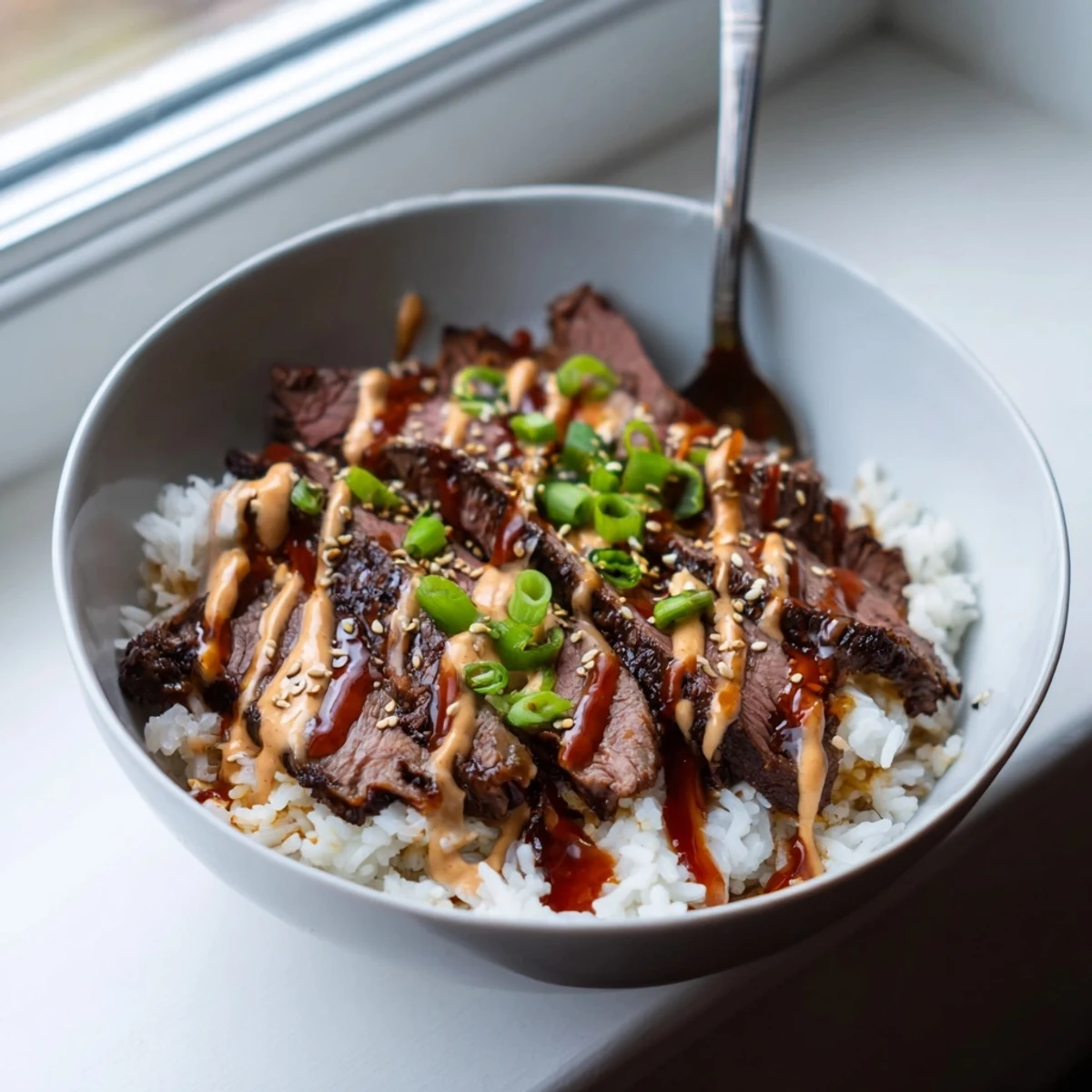 Sliced marinated steak nestled on warm rice with crisp veggies and creamy sriracha drizzle