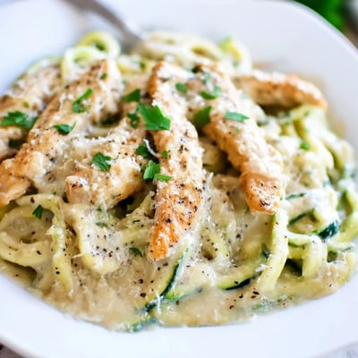 Tender chicken strips tossed with spiralized zucchini noodles in a rich, garlicky Alfredo sauce in a skillet.