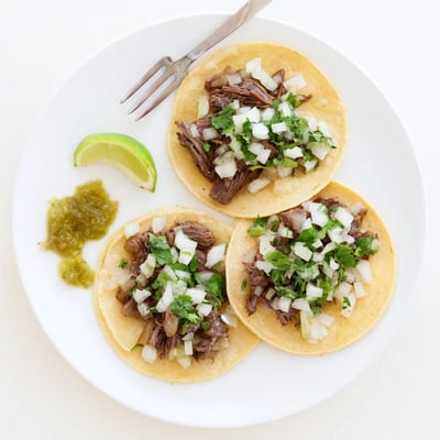 A close-up of juicy Beef Cheeks tacos garnished with lime wedges, cilantro, and onion on a rustic table.
