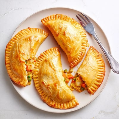 Overhead view of six hand pies filled with spiced vegetables in creamy sauce, served with a small bowl of mango chutney.