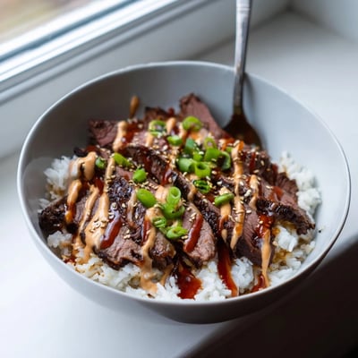 Sliced marinated steak nestled on warm rice with crisp veggies and creamy sriracha drizzle